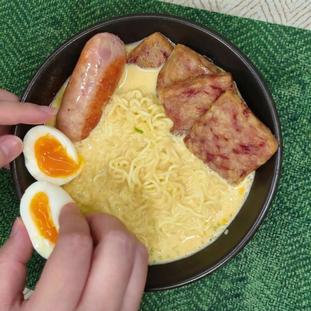 Mani che posizionano due metà di uovo barzotto con tuorlo arancione in una ciotola di ramen accanto a una salsiccia e spam.