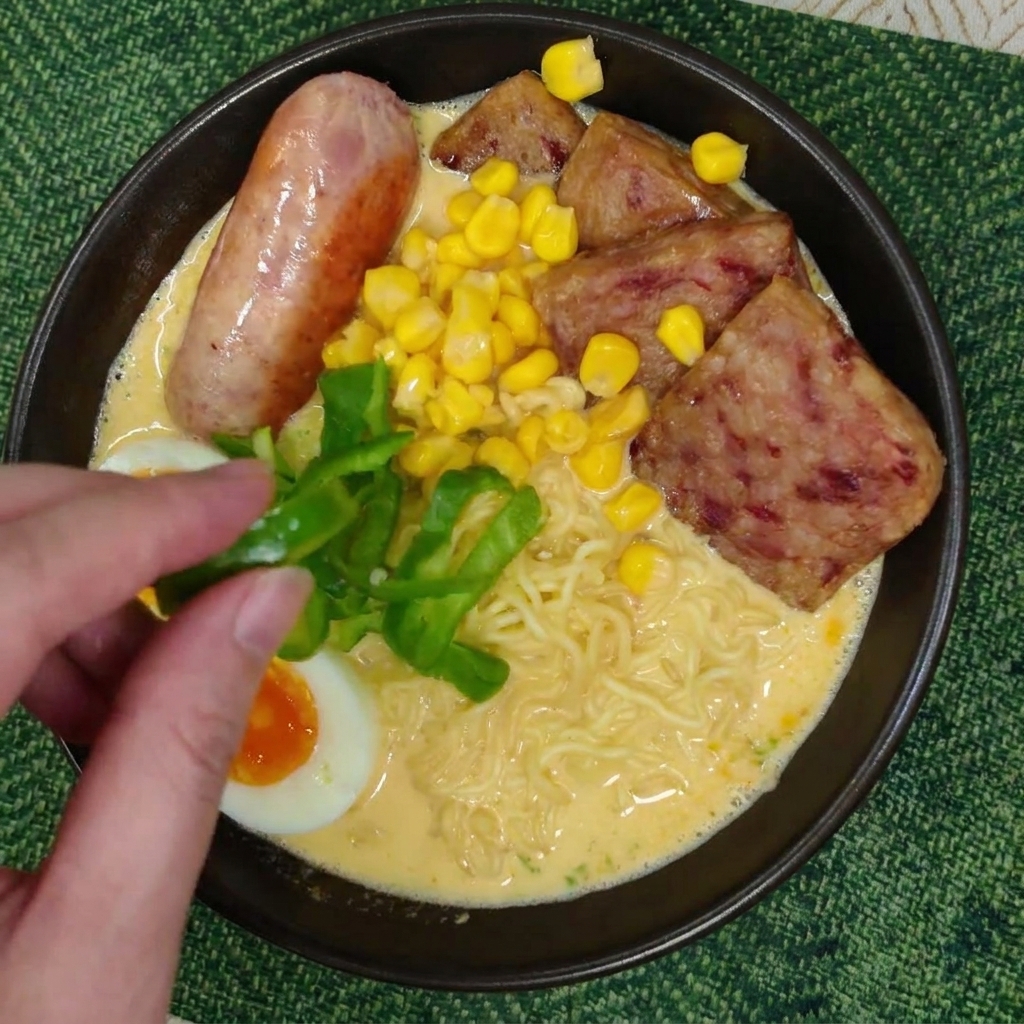 Una mano che posiziona fettine di peperone verde fresco su una ciotola di ramen guarnita.