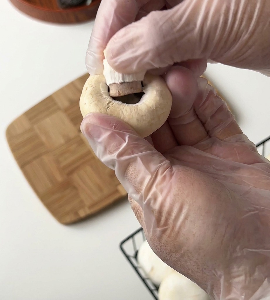 Una persona con guanti di plastica rimuove il gambo da un fungo champignon su un tagliere di legno.