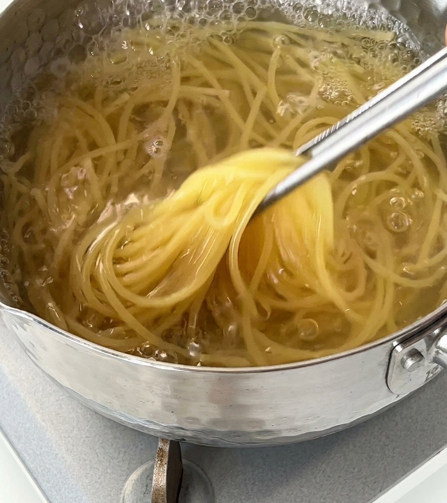 Dettaglio di spaghetti sollevati da una pentola di acqua bollente con pinze metalliche.