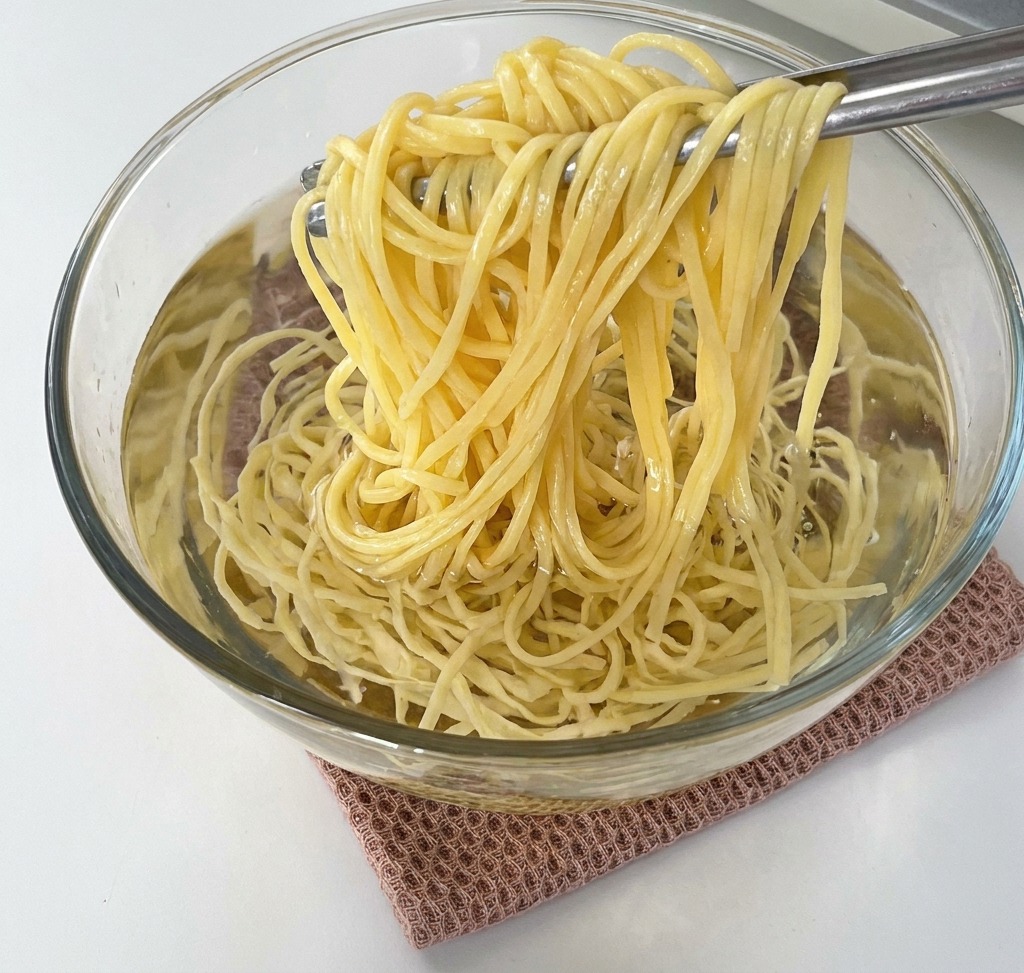 Pinze che sollevano spaghetti cotti da una ciotola di vetro piena dacqua fredda.