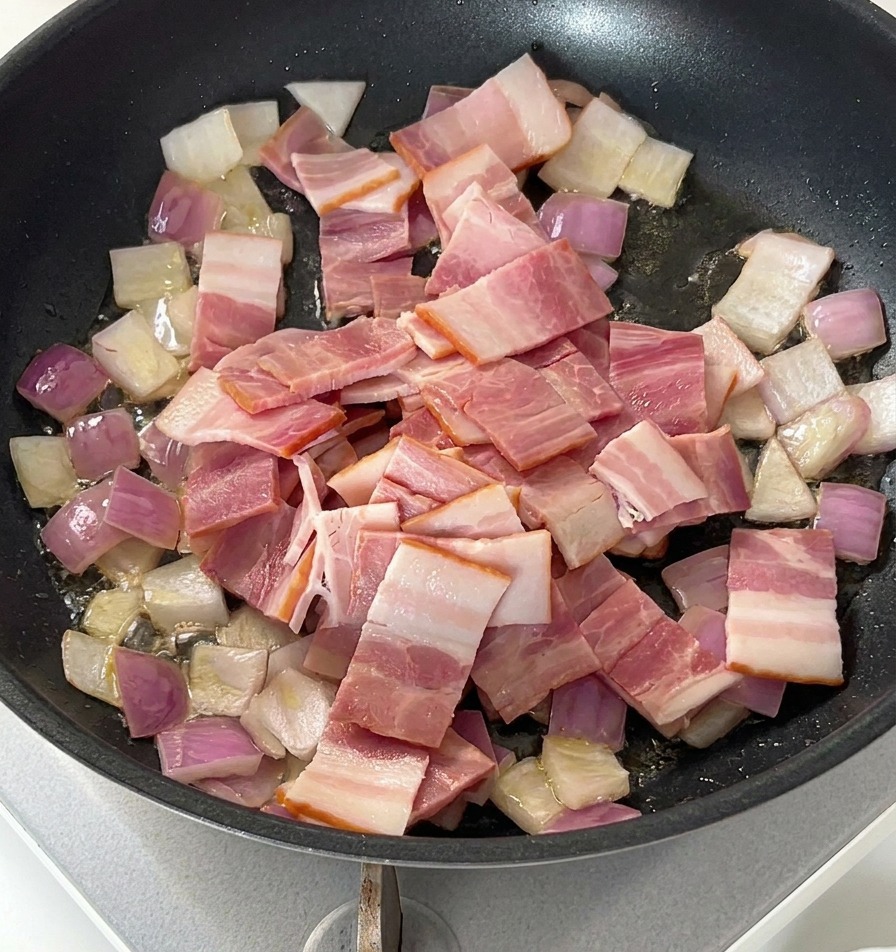 Pezzi di pancetta cruda sopra le cipolle soffritte in una padella nera.