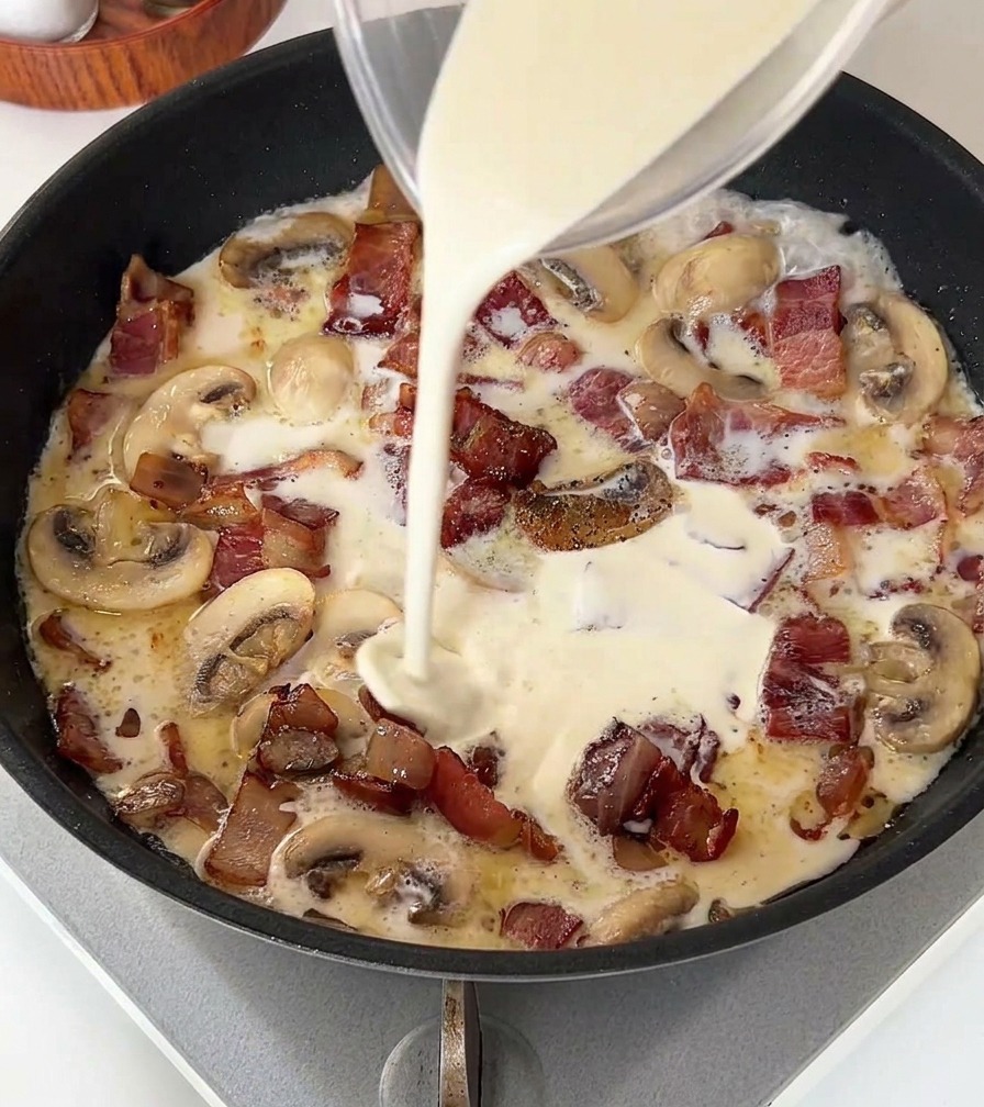 Panna liquida versata da un misurino di vetro in una padella con pancetta e funghi.