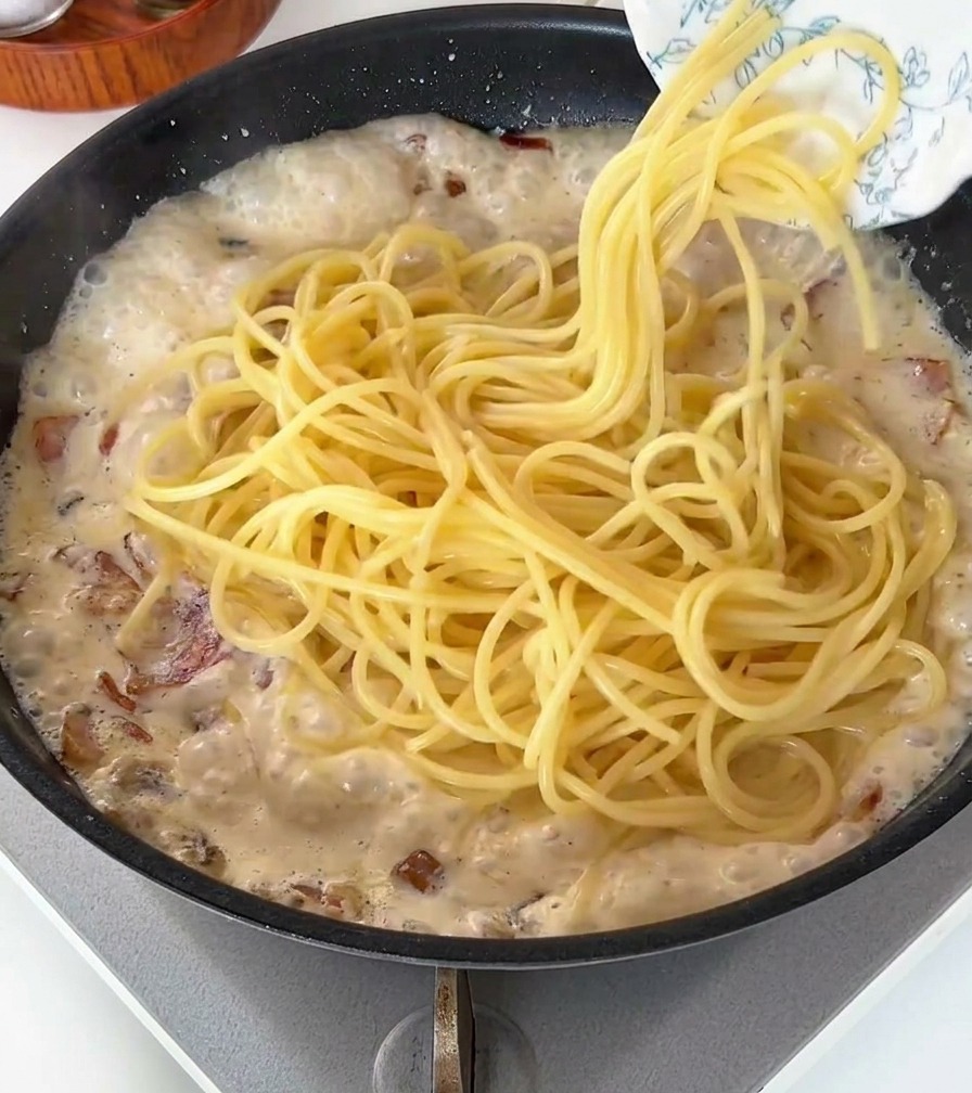 Spaghetti cotti versati in una padella con salsa cremosa bianca e pancetta.