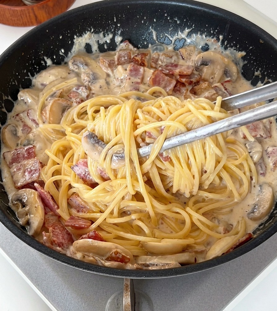 Pinze che mescolano gli spaghetti in una salsa densa e cremosa con fette di funghi e pancetta.