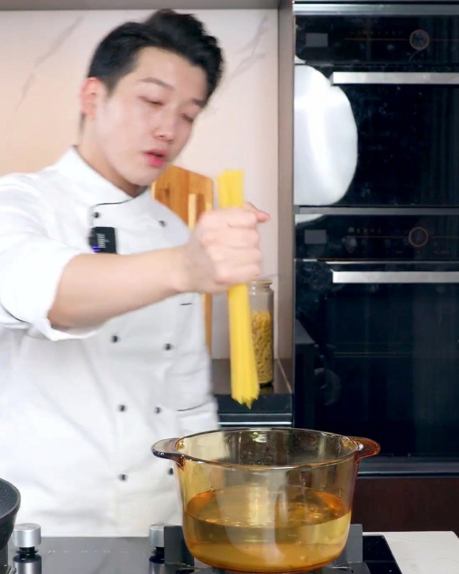 Uno chef in divisa bianca tiene un mazzo di spaghetti secchi sopra una pentola di vetro trasparente con acqua bollente.