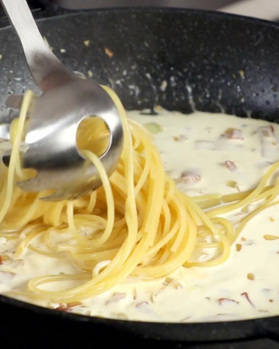 Un forchettone da pasta solleva gli spaghetti cotti e li tuffa nella salsa di panna in padella.