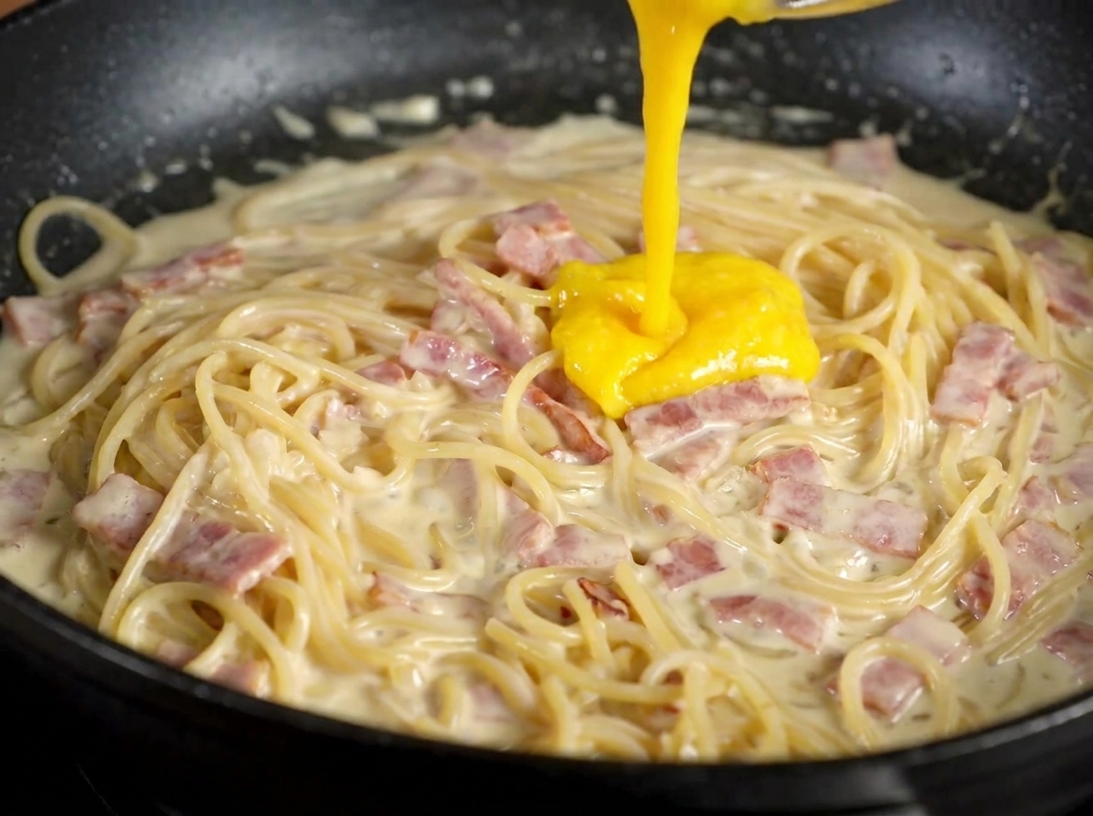 Composto di tuorlo e formaggio versato sopra la pasta cremosa con pancetta in padella.