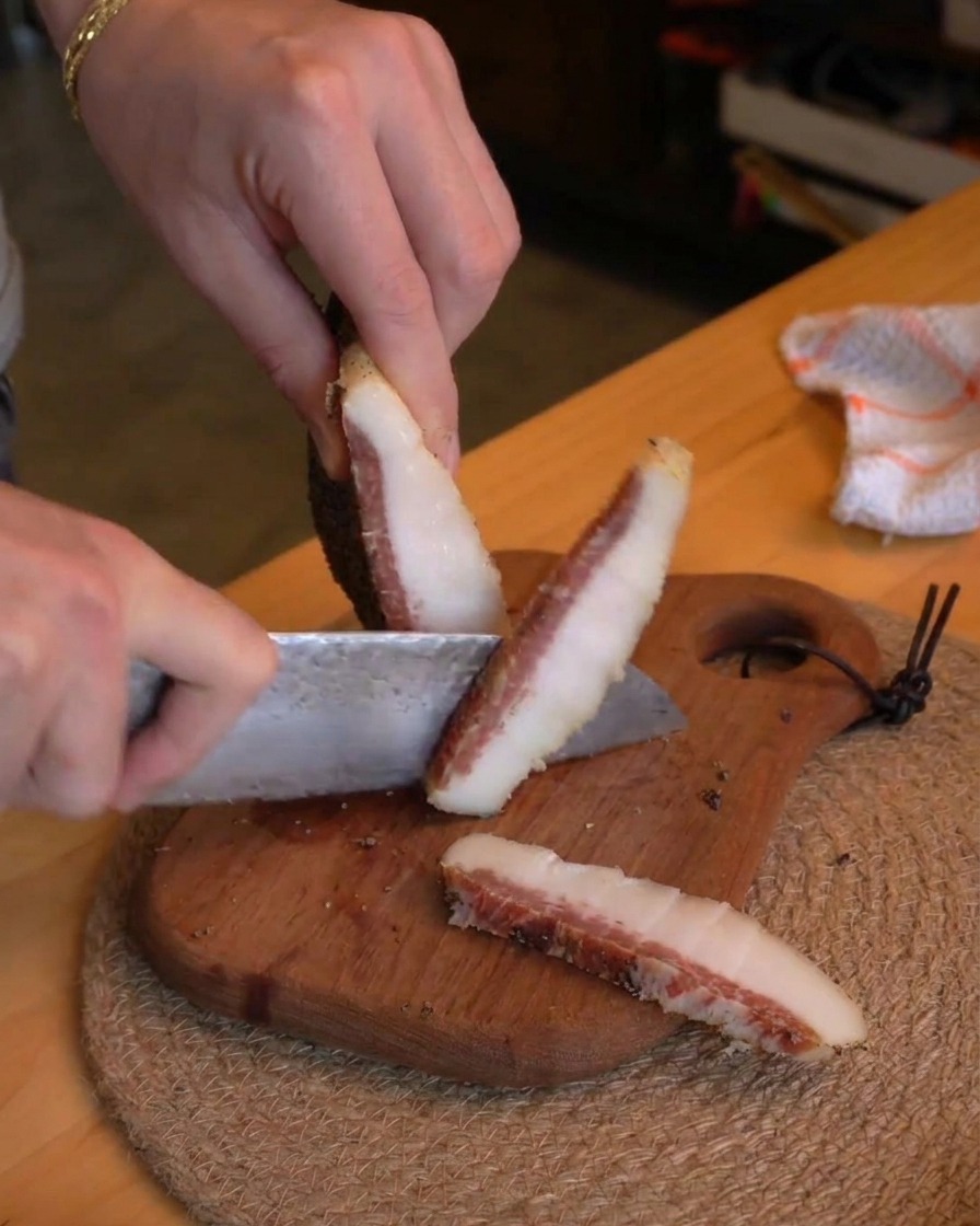 Una persona che usa un coltello da chef per tagliare spesse fette di guanciale stagionato a listarelle su un tagliere di legno.
