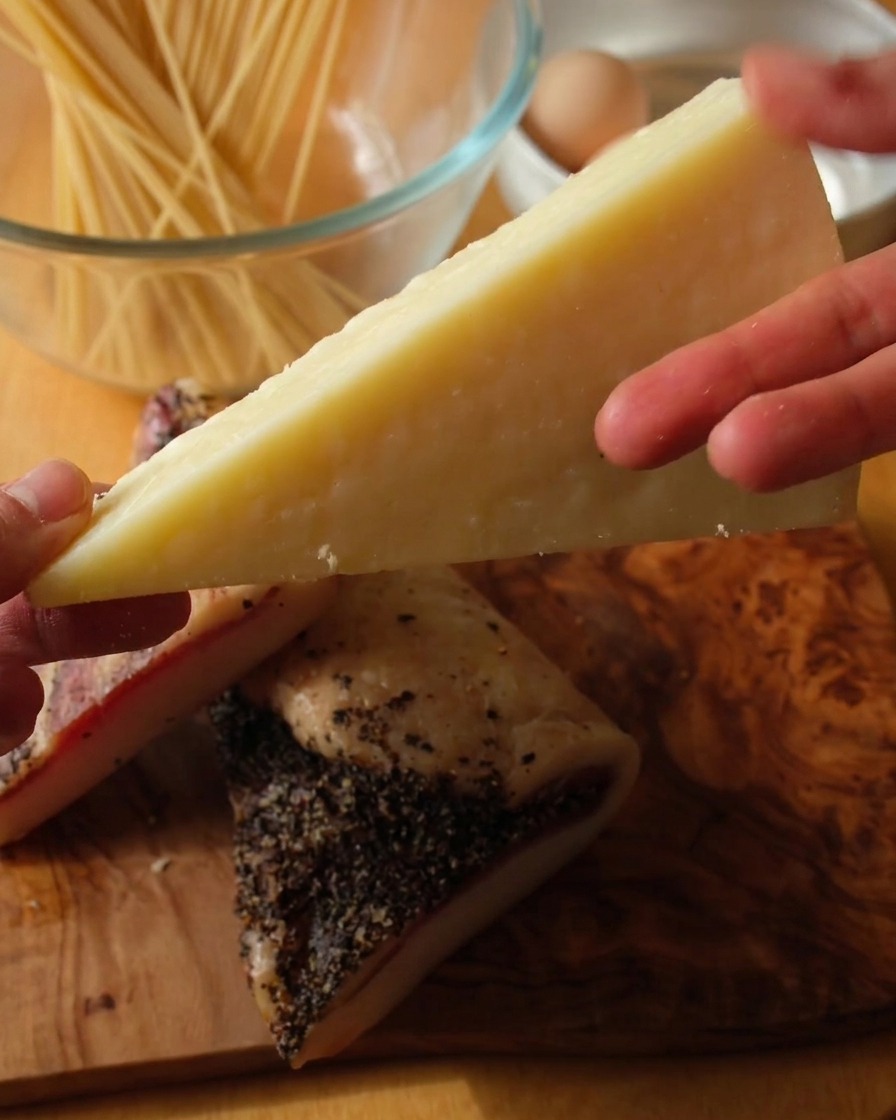 Primo piano di una mano che tiene uno spicchio di Pecorino Romano sopra due fette spesse di guanciale pepato su un piano di legno.