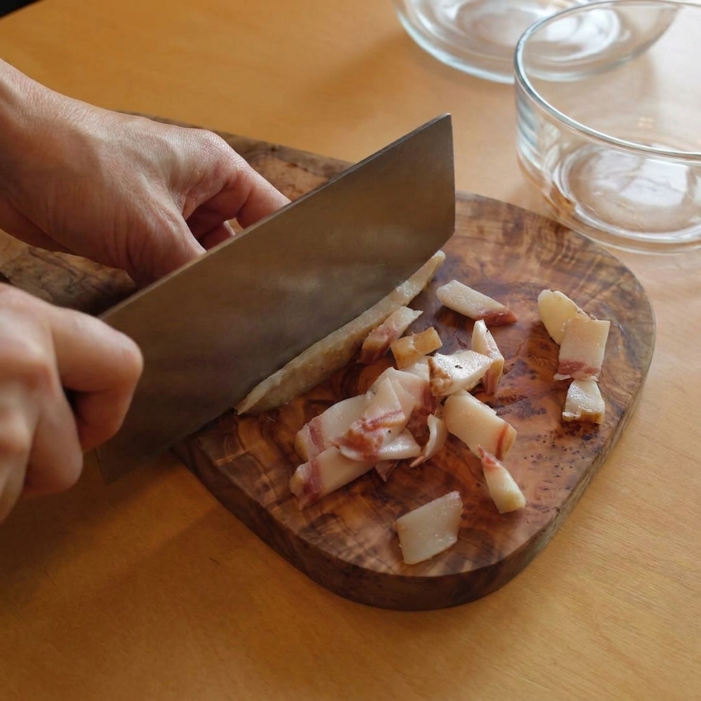 Una persona usa un coltello da cucina per tagliare a strisce il guanciale su un tagliere di legno rustico.