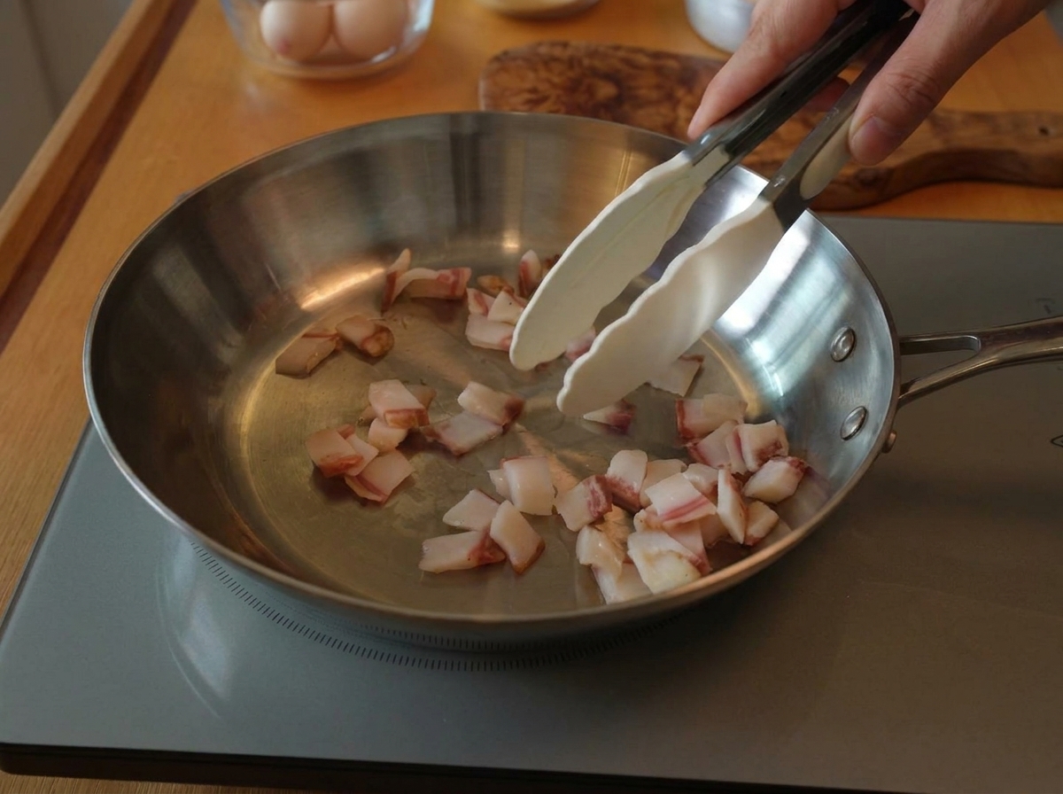 Pinze che mescolano pezzi di guanciale crudo in una padella di acciaio inossidabile su un piano a induzione.