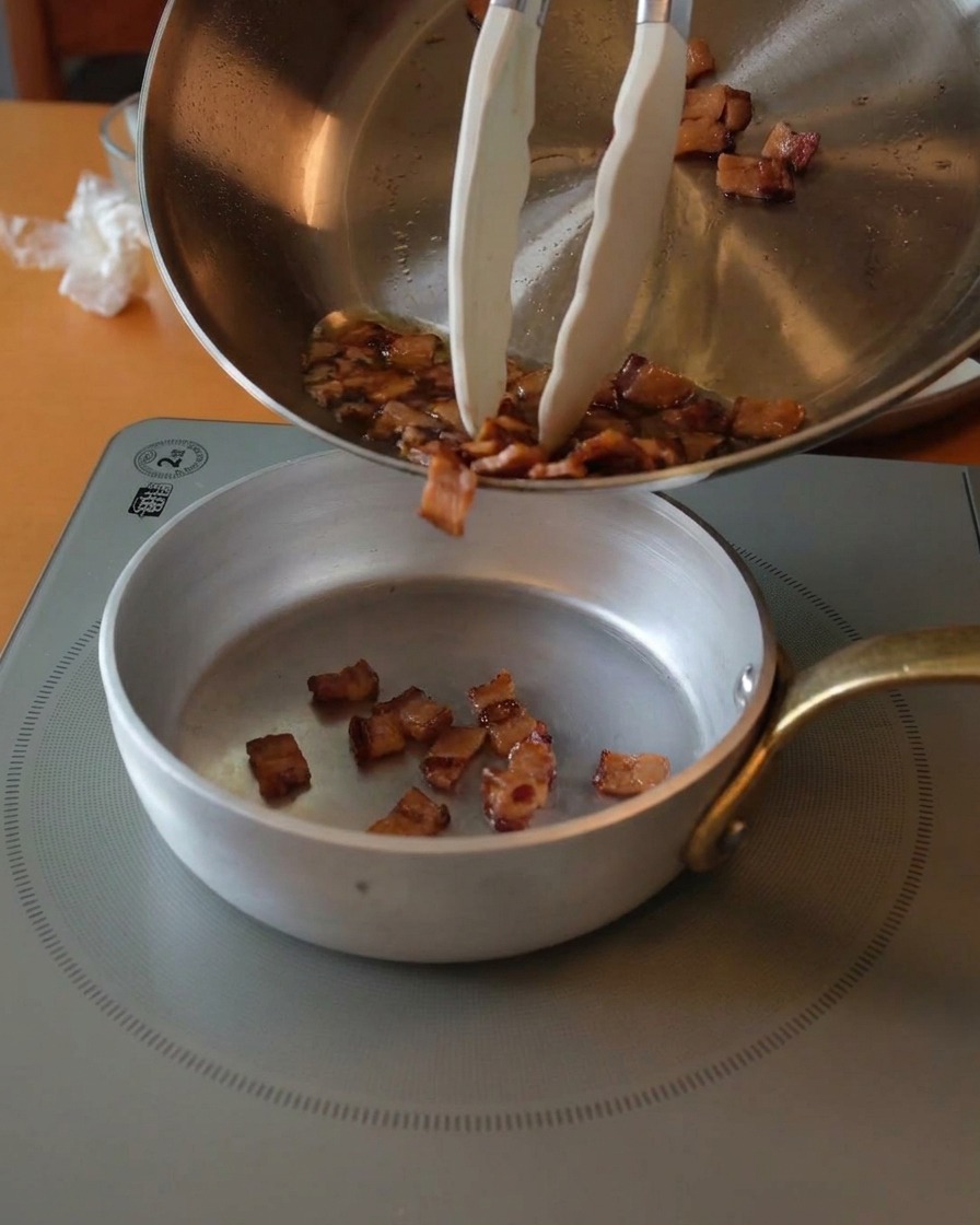 Pezzetti di guanciale dorati e croccanti versati da una padella in una ciotolina dargento usando delle pinze.
