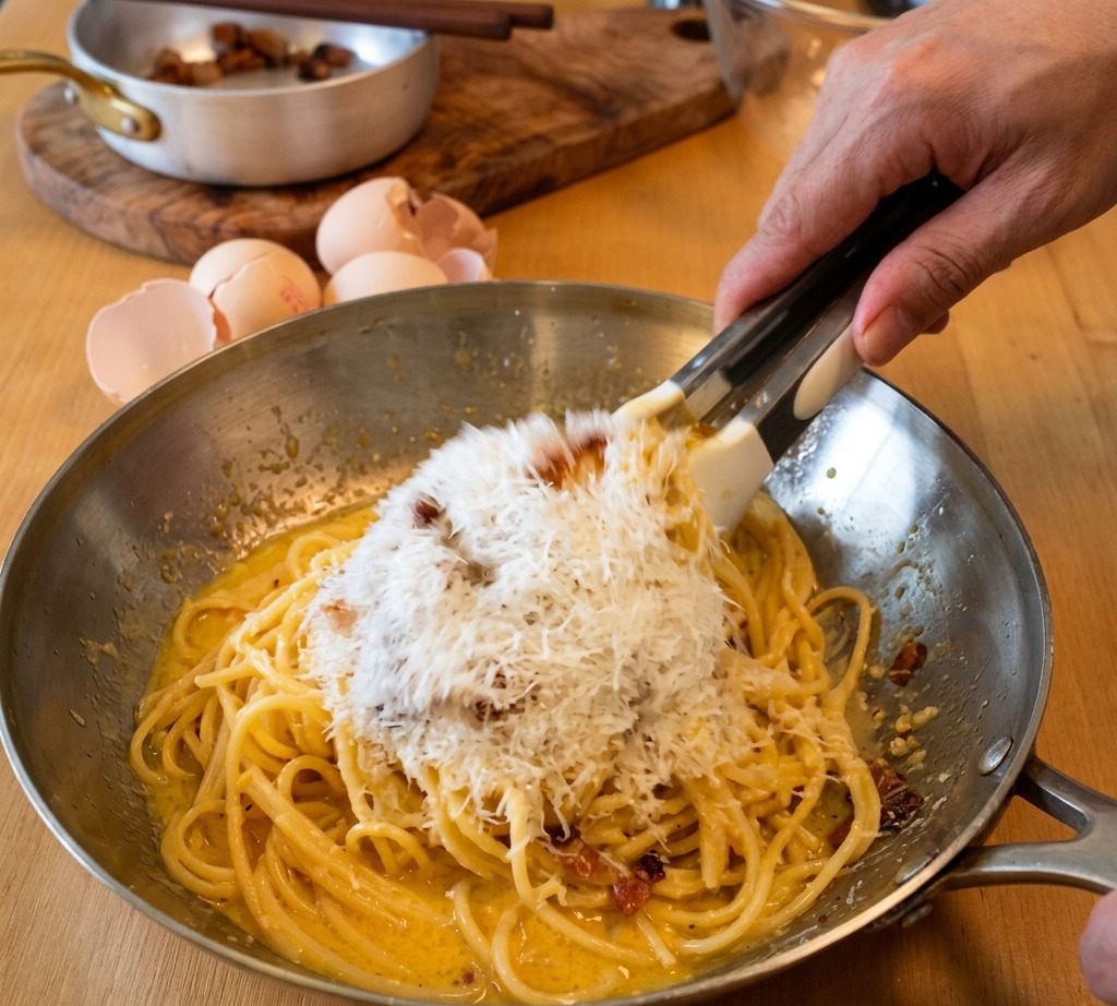 Pinze che saltano gli spaghetti in padella mentre una montagna di Pecorino grattugiato viene aggiunta alla crema duovo.