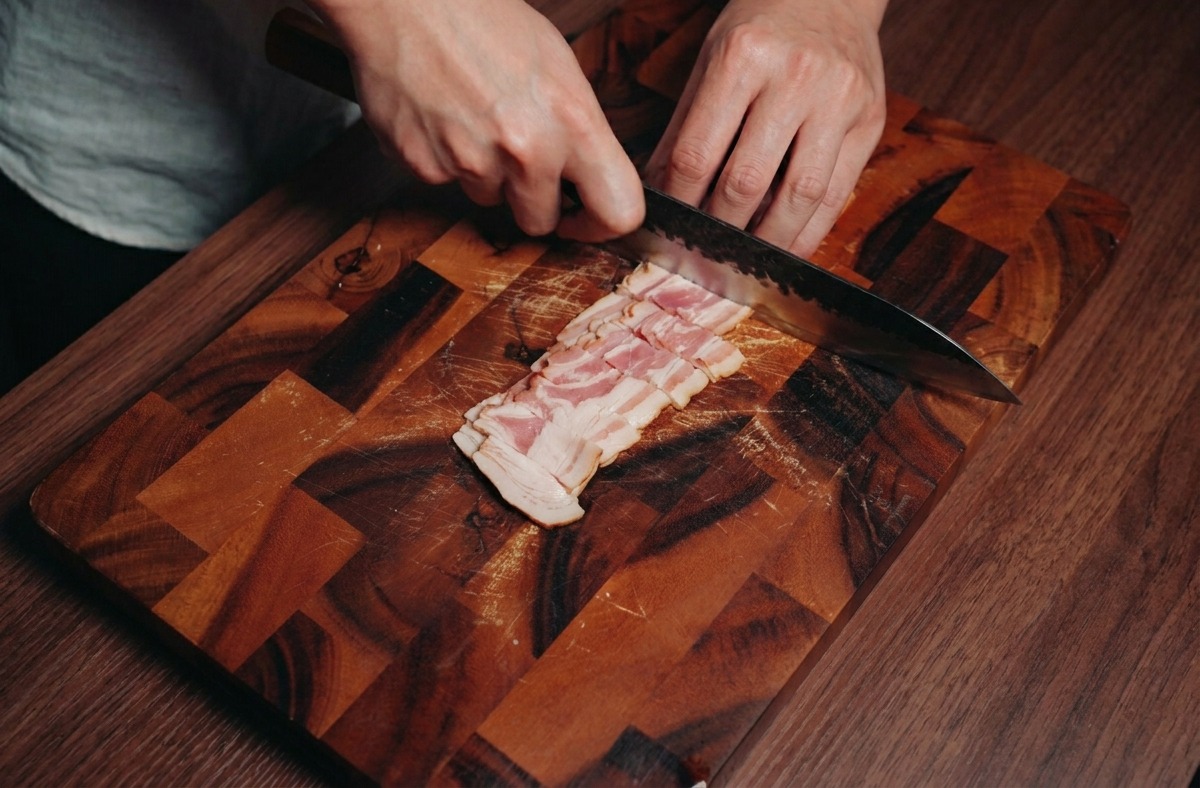 Una persona usa un coltello da chef per tagliare strisce di pancetta in piccoli cubetti su un tagliere.