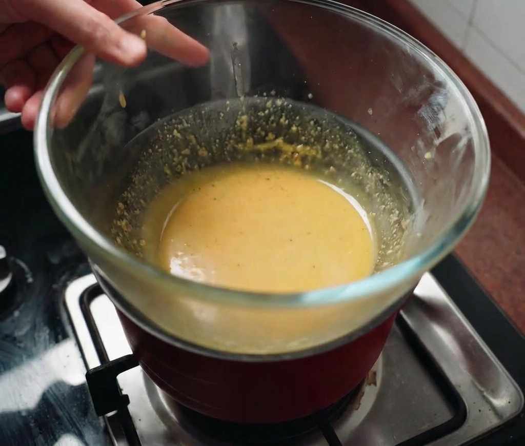Una ciotola di vetro con la salsa gialla tenuta sopra una pentola dacqua fumante.