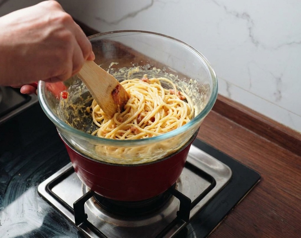 Una persona mescola pasta e pancetta nella salsa dorata dentro una ciotola sopra il vapore.