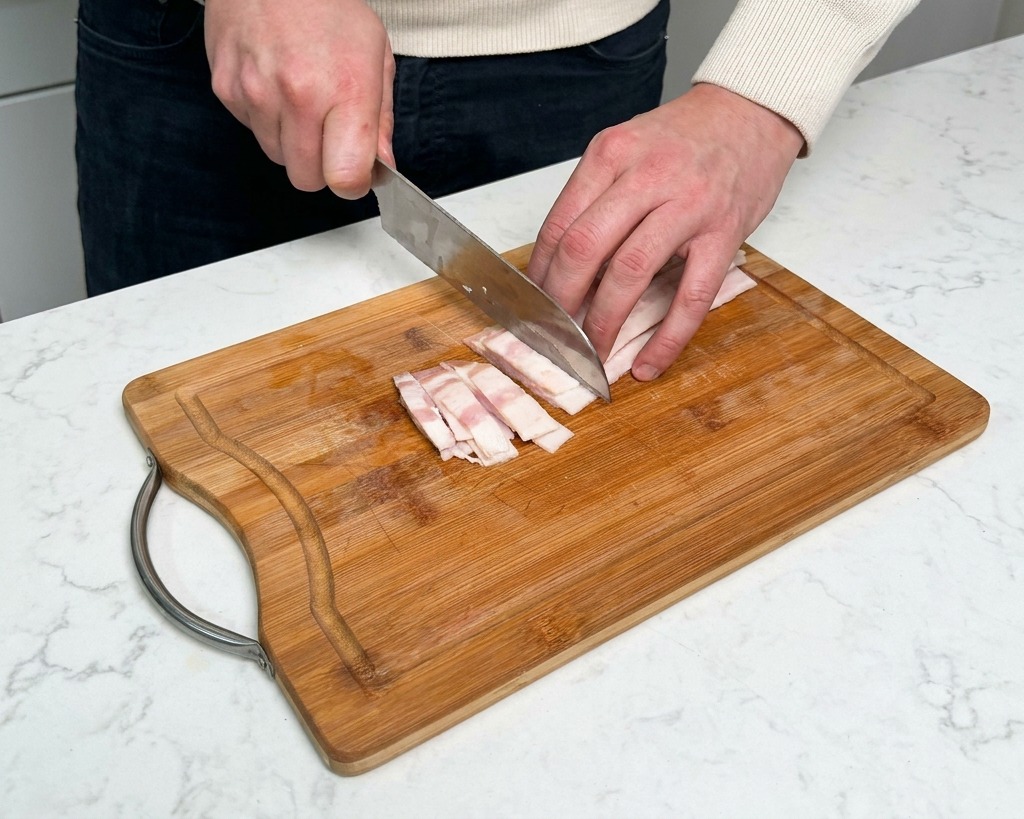 Mano che affetta strisce di pancetta cruda in piccoli rettangoli su un tagliere di legno.