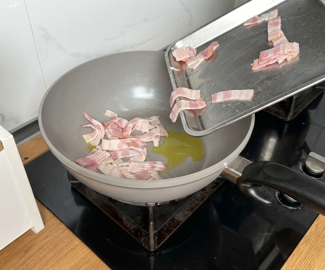 Pezzetti di pancetta versati da un vassoio di metallo in una padella capiente con olio caldo.