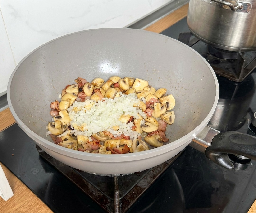 Cipolla tritata posta al centro di una padella con pancetta e funghi rosolati.