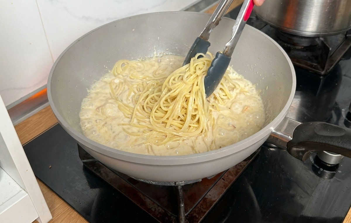 Pasta lunga cotta trasferita con pinze di metallo da una pentola alla padella con la salsa cremosa.