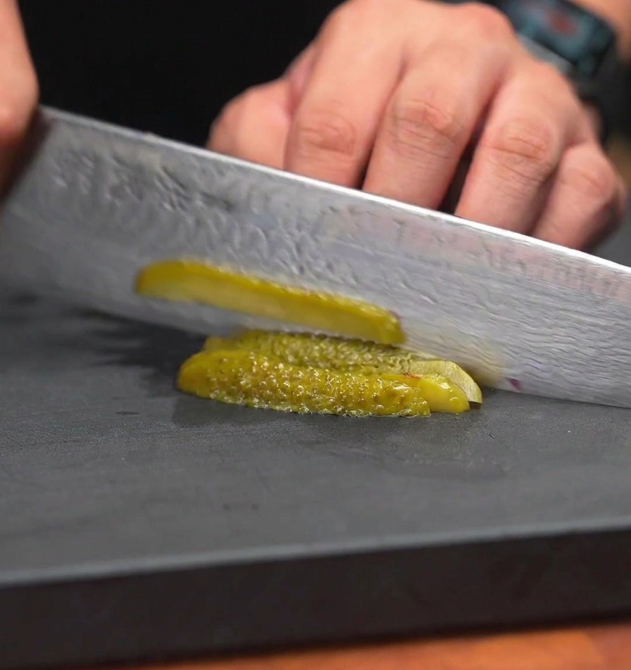 Un coltello da chef che trita cetriolini sottaceto in pezzi molto piccoli e uniformi su una superficie di lavoro nera.