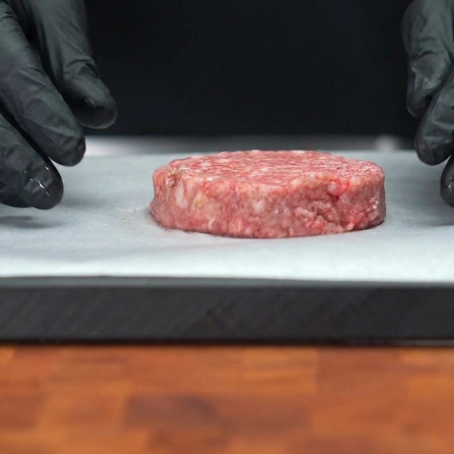 Un patty di carne di manzo crudo appoggiato su carta forno con grani visibili di pepe nero sulla superficie.