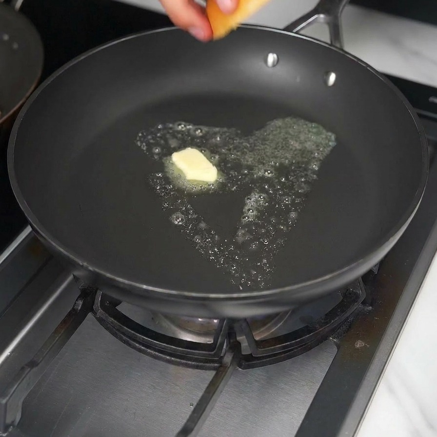 Una mano che mette un panino brioche in una padella con un cubetto di burro giallo che si scioglie.