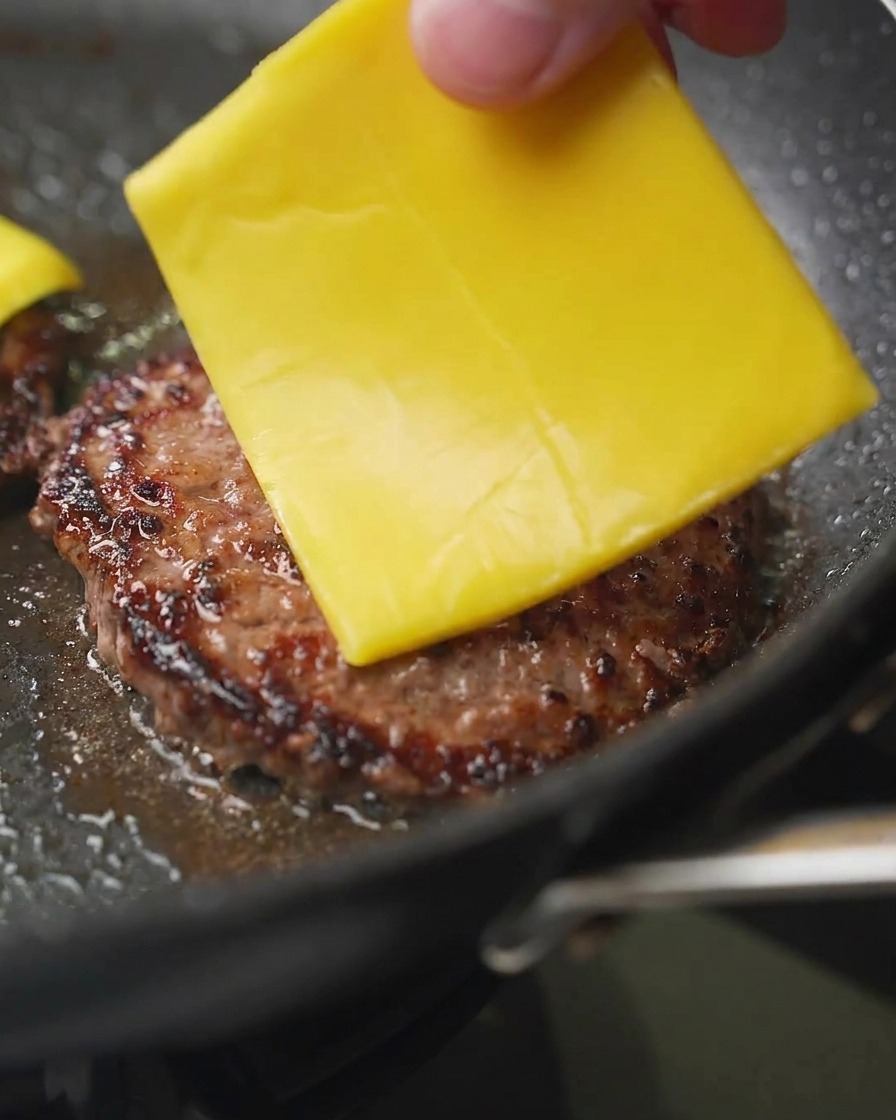 Primo piano di una mano che posiziona una fetta quadrata di cheddar giallo su un patty di manzo ben rosolato in padella.