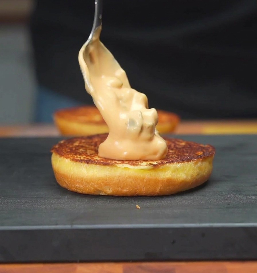 Un cucchiaio che spalma una salsa cremosa arancione sulla superficie tostata di un panino da hamburger.