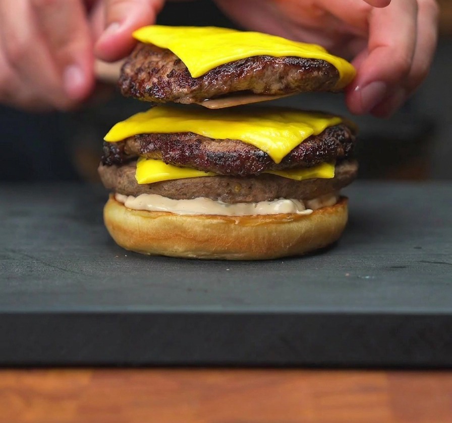 Una mano che adagia due patty di cheeseburger con formaggio fuso sopra un panino già condito con la salsa.