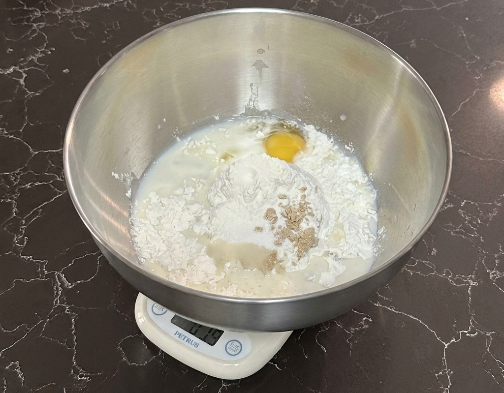 Una ciotola in acciaio su una bilancia digitale contenente farina, latte, un uovo e lievito.