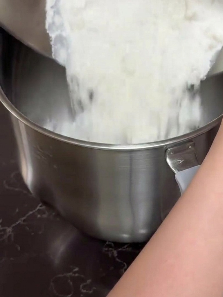 Mani che versano il composto di farina e latte da una ciotola dacciaio nel contenitore di una planetaria.