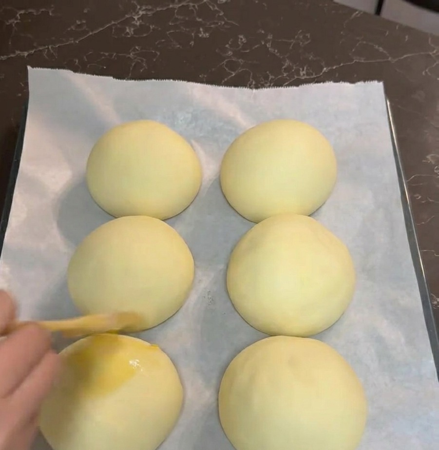 Una mano che usa un pennello da cucina per ricoprire sei palline di impasto lievitate con un uovo sbattuto su una teglia.