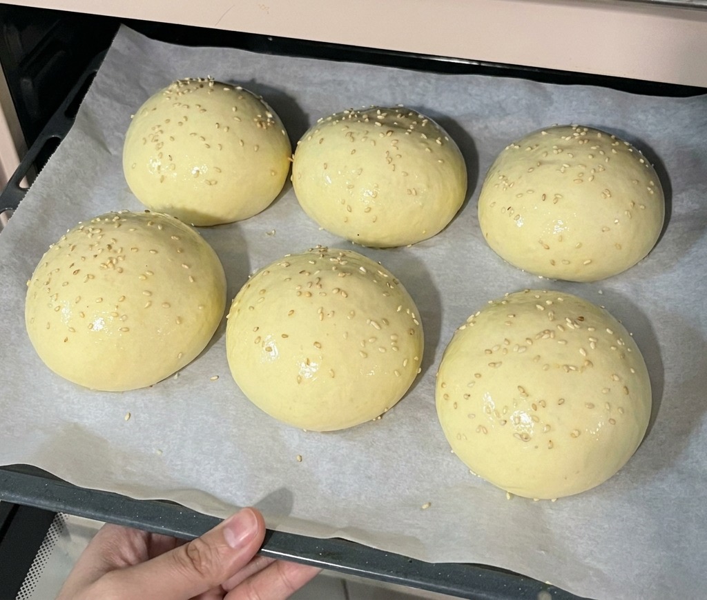 Una teglia con sei panini lievitati ricoperti di semi di sesamo che viene inserita in un forno preriscaldato.