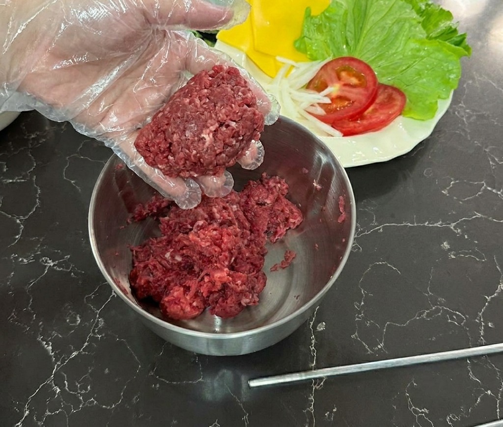 Una mano con un guanto di plastica trasparente che tiene una pallina di carne macinata cruda sopra una ciotola.
