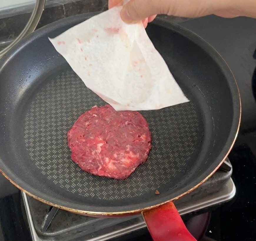 Un burger di manzo crudo inserito in una padella calda mentre la carta forno viene rimossa a mano.
