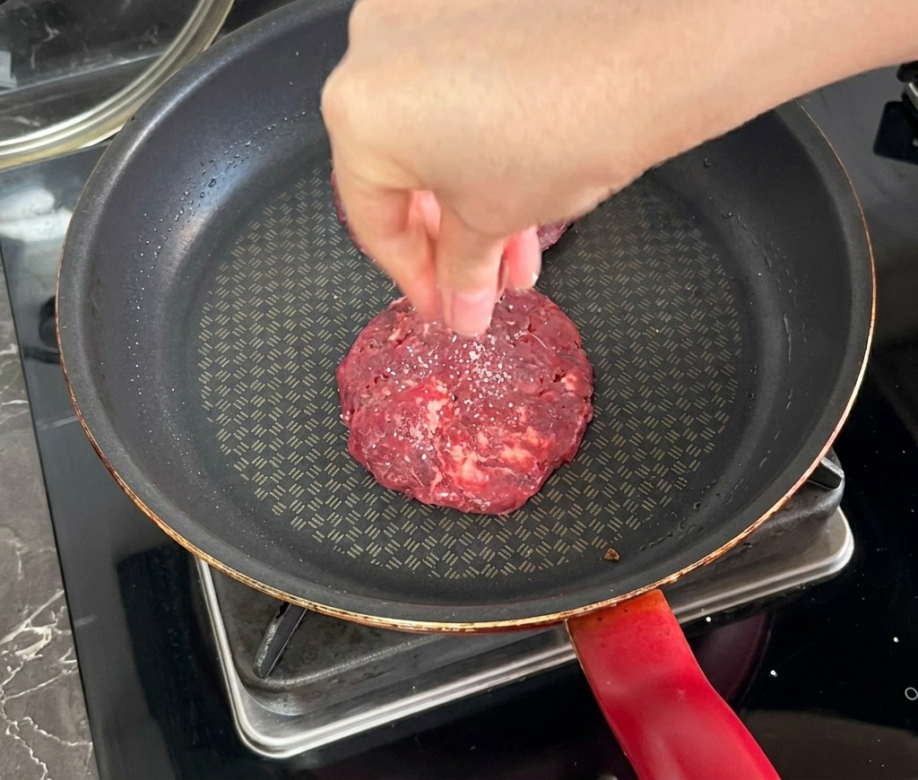 Una mano che sparge il sale su un burger di manzo che sta cuocendo in una padella antiaderente scura.