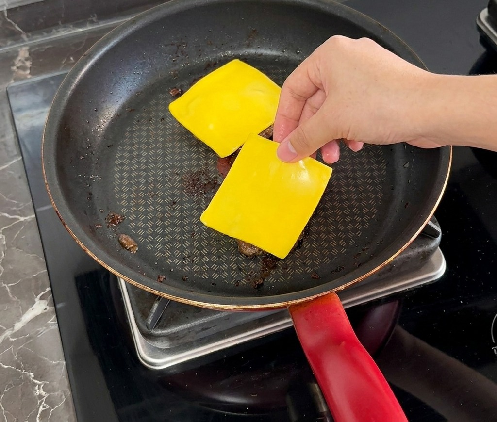 Una mano che mette una fetta quadrata di formaggio giallo su un hamburger cotto in padella.