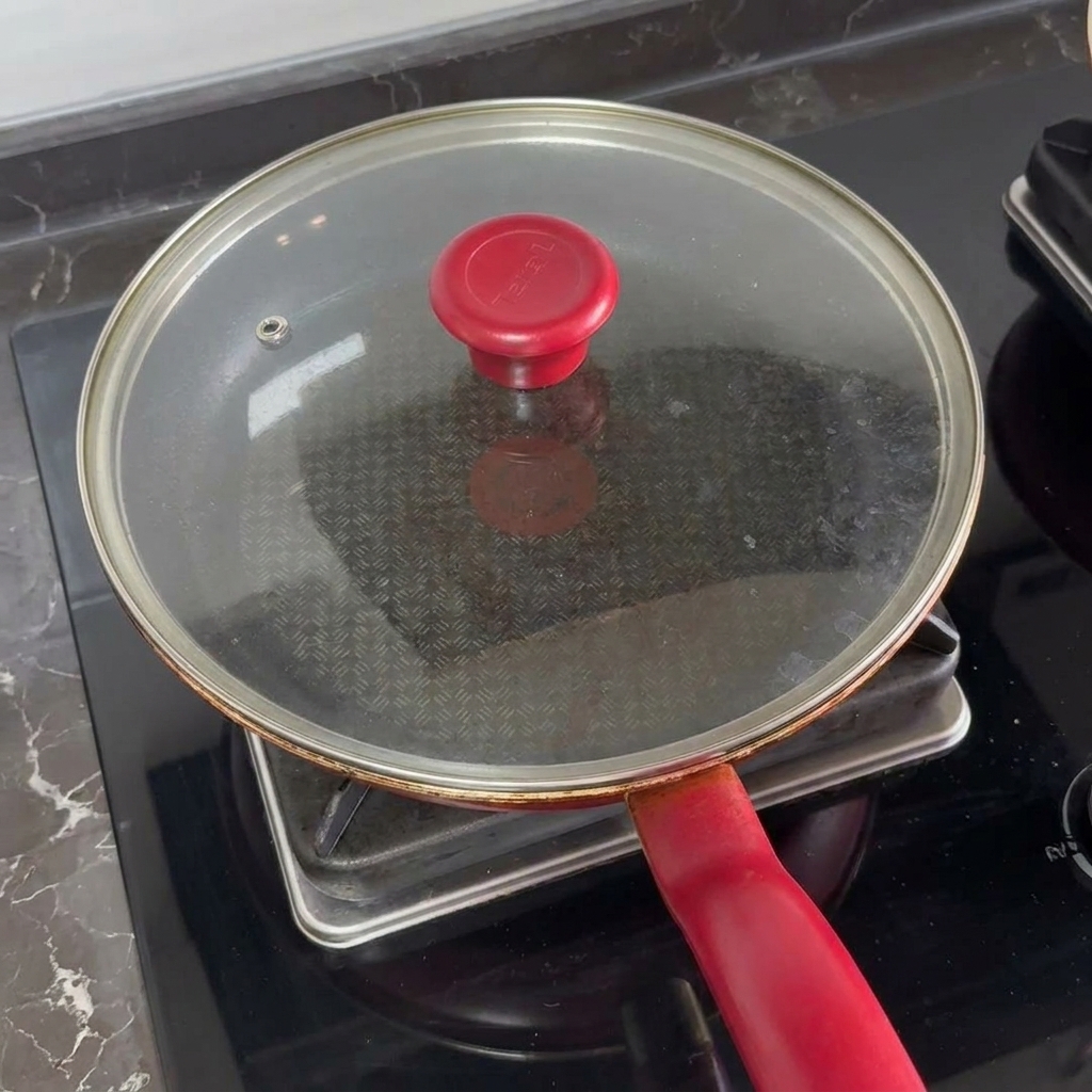 Un coperchio di vetro con manico rosso che copre una padella sui fornelli per fondere il formaggio sopra i burger.