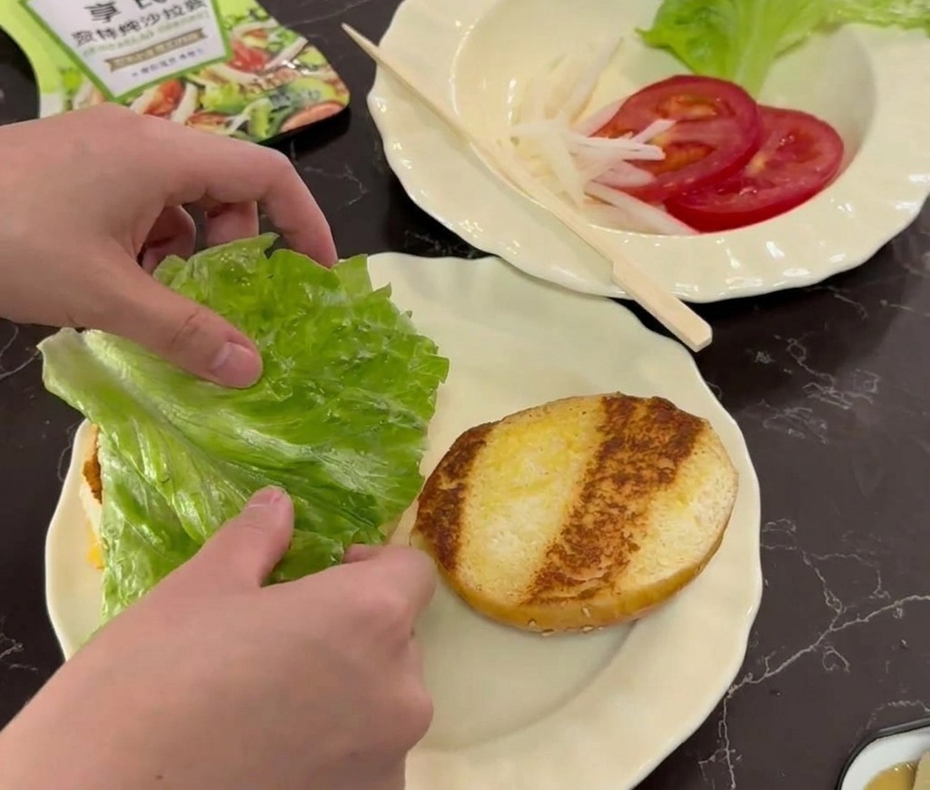 Mani che posizionano una foglia di lattuga fresca su un panino tostato in un piatto bianco decorato.