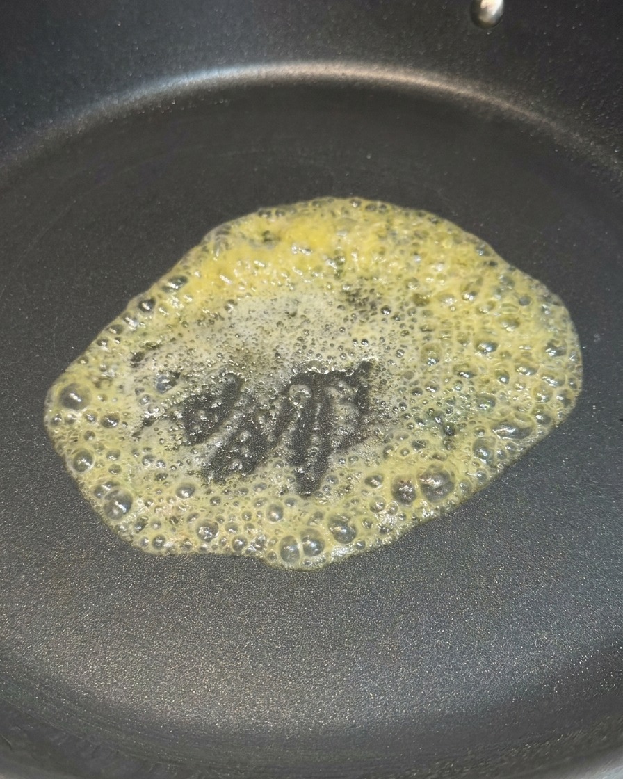 Una noce di burro giallo che si scioglie e sfrigola al centro di una padella antiaderente nera.