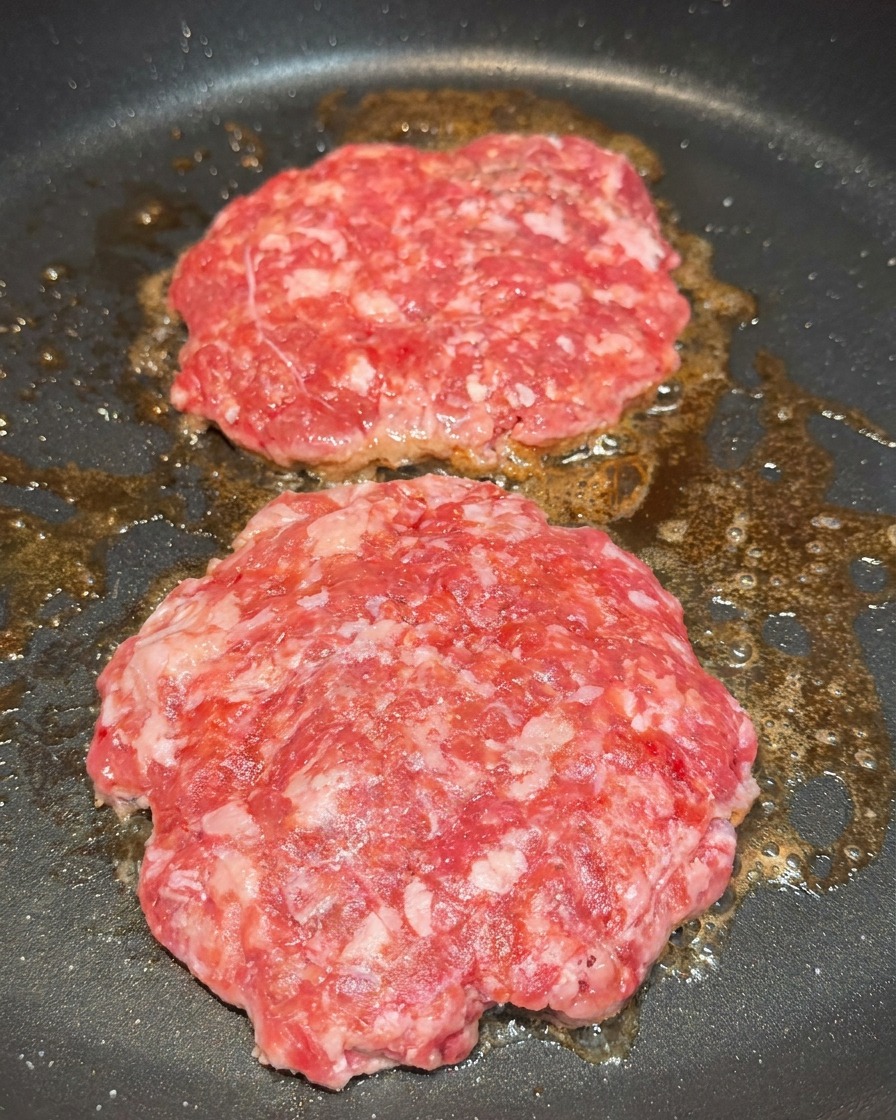 Due hamburger in padella conditi con grani visibili di sale e pepe nero.