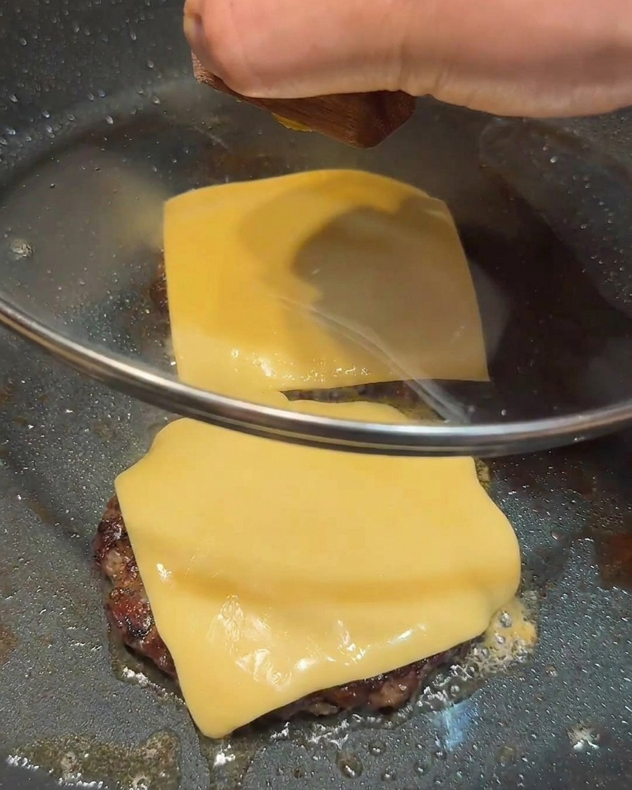 Un coperchio di vetro che copre una padella con due hamburger sormontati da formaggio fuso.