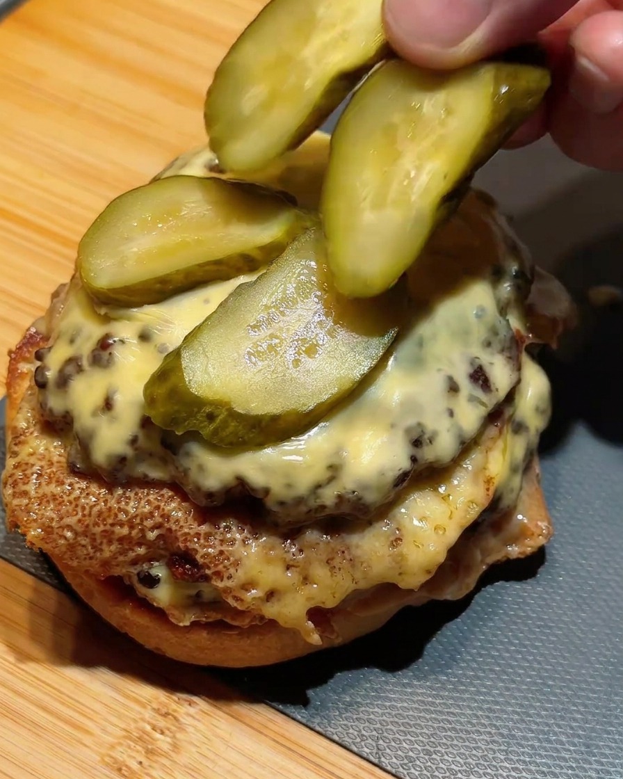 Dita che dispongono fette di cetriolini verdi sopra il formaggio fuso dellhamburger superiore.