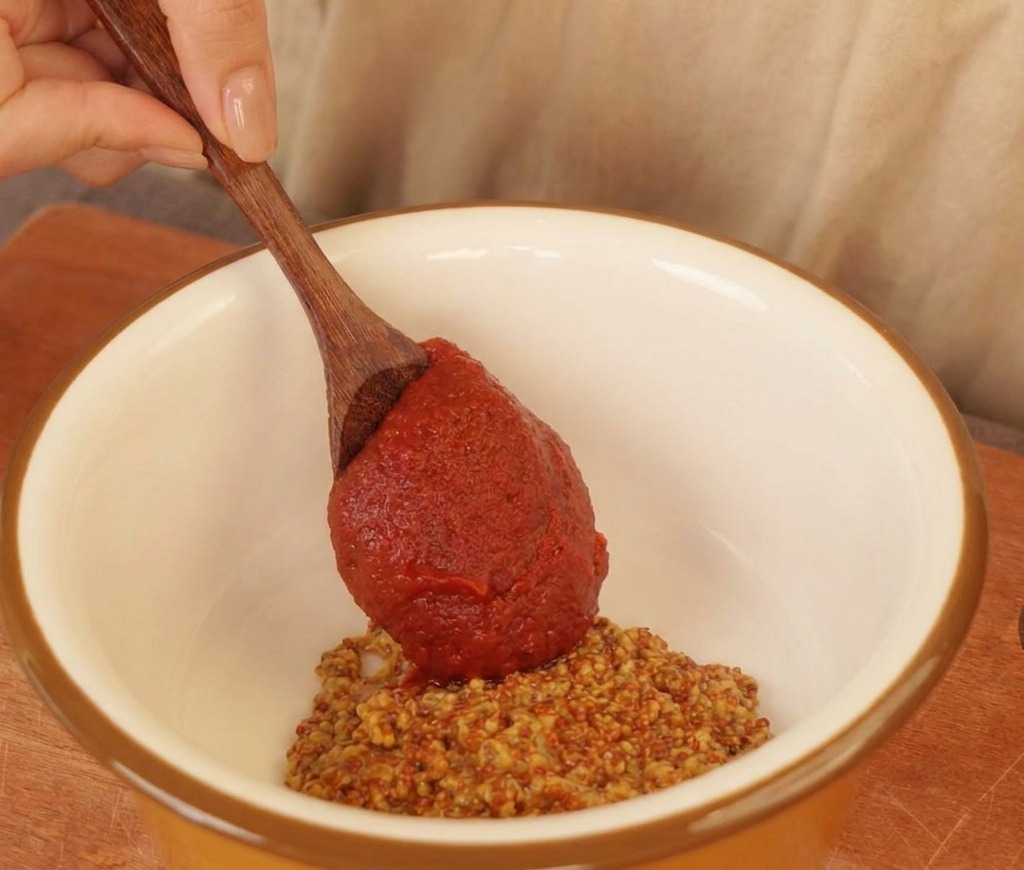 Un cucchiaio di legno che aggiunge salsa al peperoncino in una ciotola con senape in grani e maionese.
