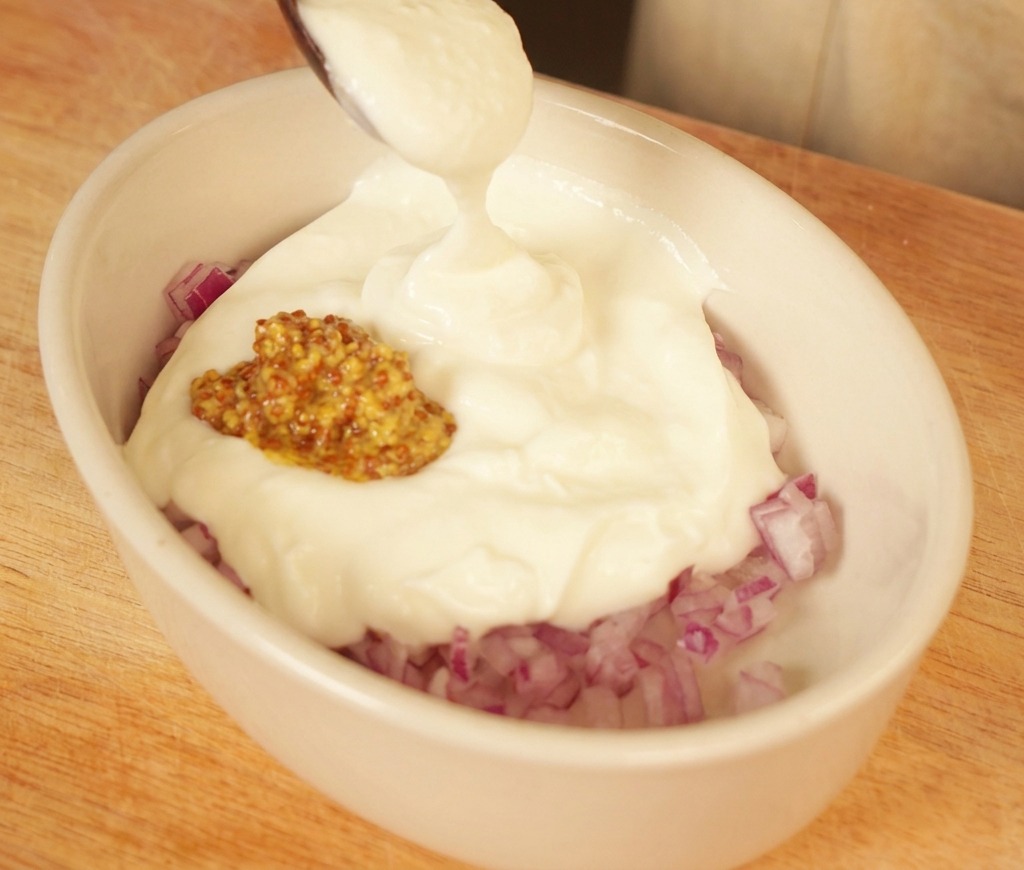 Un cucchiaio di yogurt bianco cremoso aggiunto a un piattino con cipolle rosse tritate e senape.