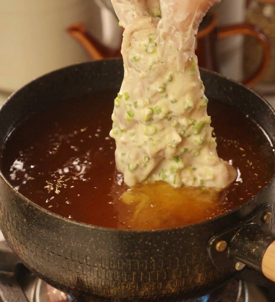 Una mano che immerge un pezzo di pollo ricoperto di pastella al cipollotto in una pentola di olio bollente.