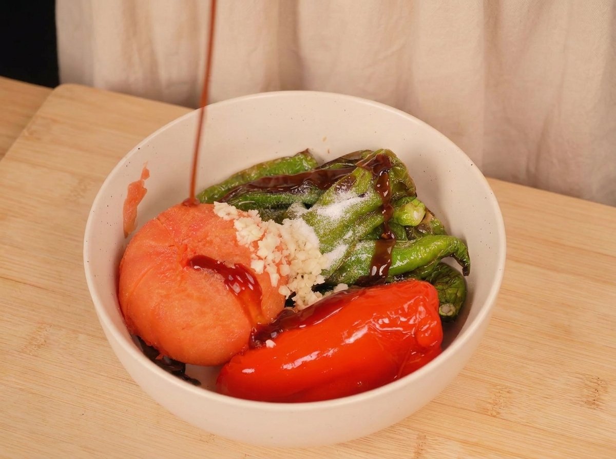 Salsa dostriche scura versata sopra peperoni arrostiti, un pomodoro, aglio tritato e sale in una ciotola bianca.