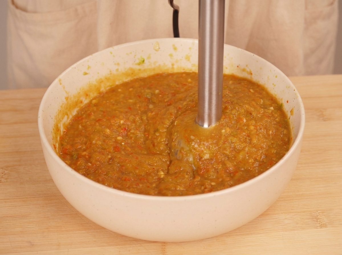 Un frullatore a immersione utilizzato allinterno di una ciotola bianca per trasformare le verdure arrosto in una salsa densa arancione-verde.